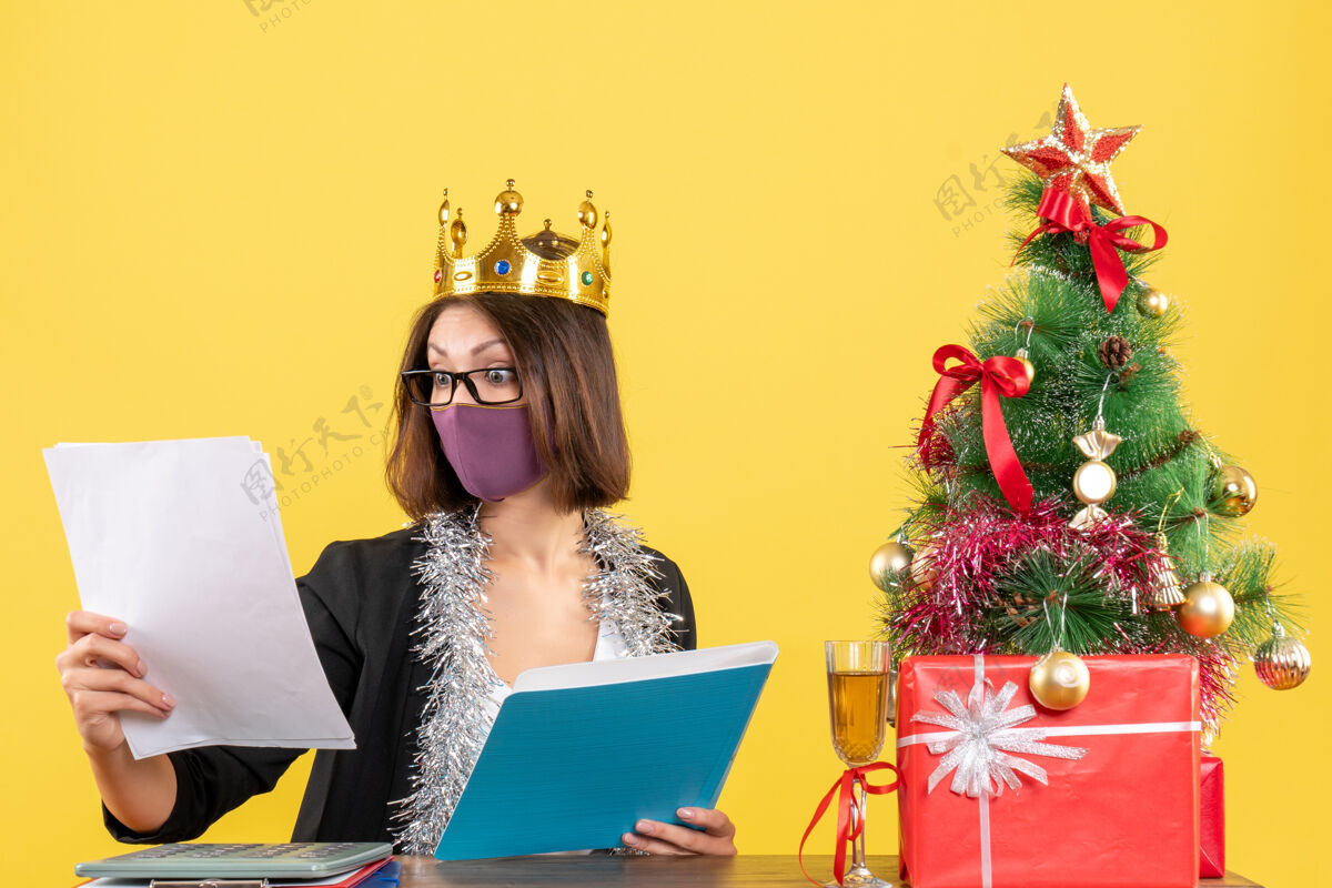 美女Xsmas心情与穿着西装戴着皇冠戴着医用口罩的美丽女士在黄色办公室查看文件人笔记本电脑穿着