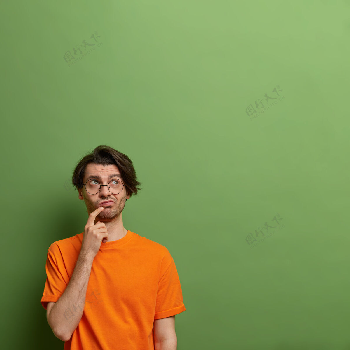 选择沉思的优柔寡断的成年男子向上看 把手指放在嘴边 穿着休闲的橙色t恤 对着绿墙摆姿势 留着复制空间 为你的晋升做准备思考思想眼镜