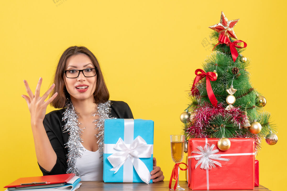情感情绪化的商务女士 穿着西装 戴着眼镜 手里拿着礼物 坐在一张桌子旁 桌子上放着圣诞树抱着快乐眼镜