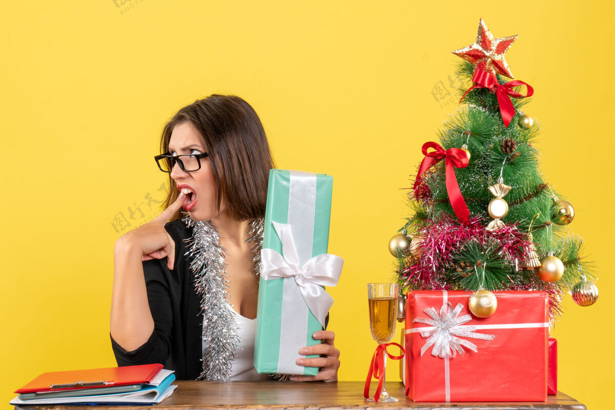 快乐困惑的商务女士穿着西装 戴着眼镜 惊讶地展示着她的礼物 坐在一张桌子旁 桌子上放着一棵圣诞树眼镜漂亮肖像