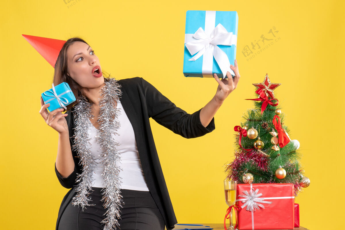 美丽的女士圣诞心情惊喜美丽的女士戴着圣诞帽拿着礼物在黄色的办公室里模特心情女士