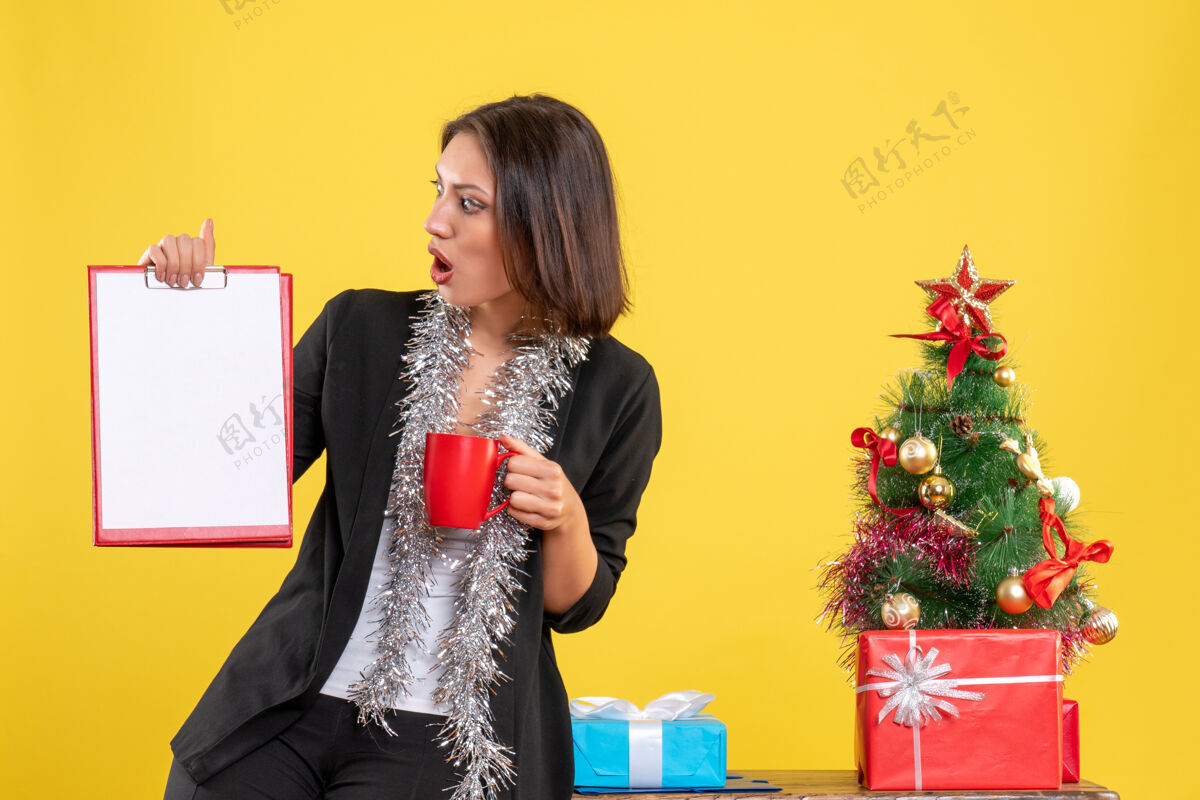 头发圣诞节的心情与惊喜美丽的女士站在办公室和举行文件杯在黄色的办公室心情肖像花束