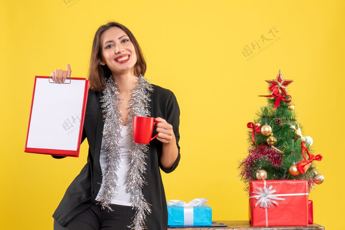 女士圣诞节的心情 微笑的美丽女士站在办公室里 拿着文件杯在办公室里的黄色肖像圣诞节美丽的女士