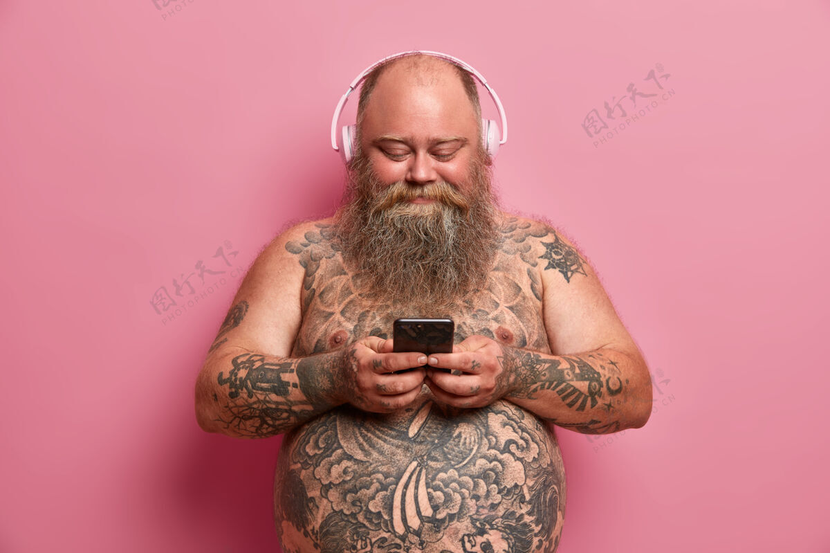 超重身材丰满 赤裸着发髻 大肚子 戴着耳机听音乐 拿着手机 在播放列表中下载歌曲 隔离在粉红色的墙上人 超重 爱好的概念耳机音乐腹部