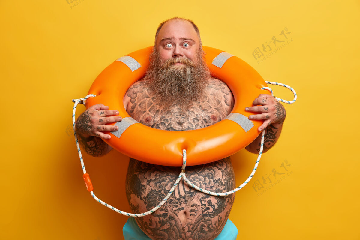 帅气夏季休息和安全游泳的概念震惊的胡子粗壮的男人赤裸着站着 有纹身的身体和大肚子 摆着充气的救生圈 等待假期 隔离在黄色的墙壁上人肥胖赤膊