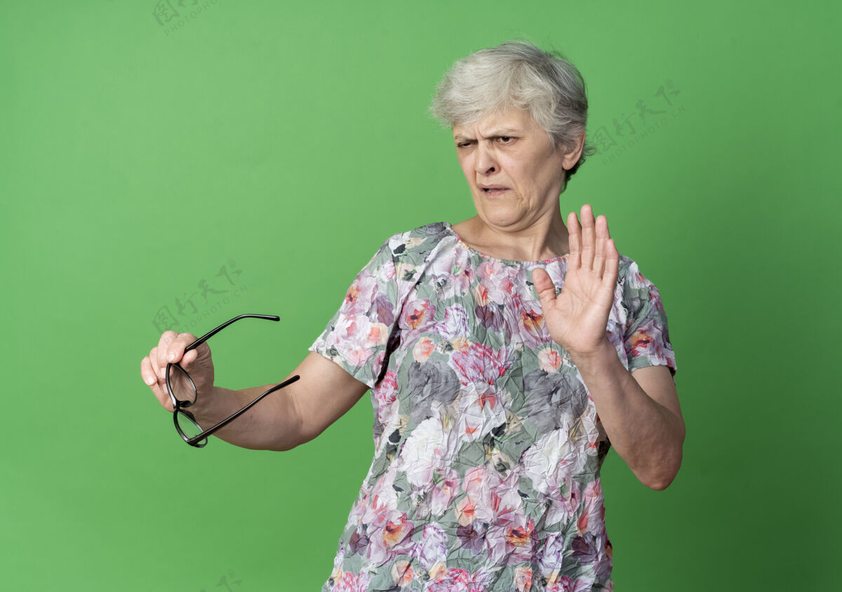 老人恼怒的老妇人举着眼镜举起手隔离在绿墙上举行手眼镜