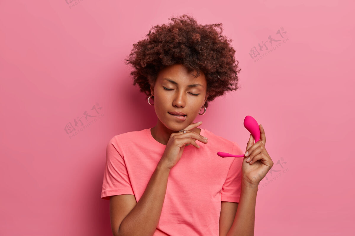 女人快乐的黑皮肤女人拿着震动器性玩具和应用程序来控制震动 咬嘴唇和满意地闭上眼睛 拿着硅胶智能震动器蛋隔离在粉红色的墙壁上情色的乐趣卷发前戏姿势