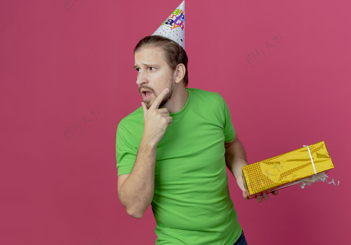 壁板戴着生日帽的迷茫帅哥拿着礼盒看着粉色墙上孤立的一面礼物帽子迷茫