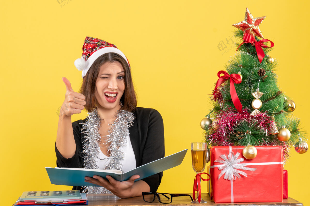 圣诞老人穿着西装 戴着圣诞老人帽和新年装饰品的商务女士正在检查文件 做着“ok”的手势 坐在一张桌子旁 桌子上放着圣诞树文件人制造