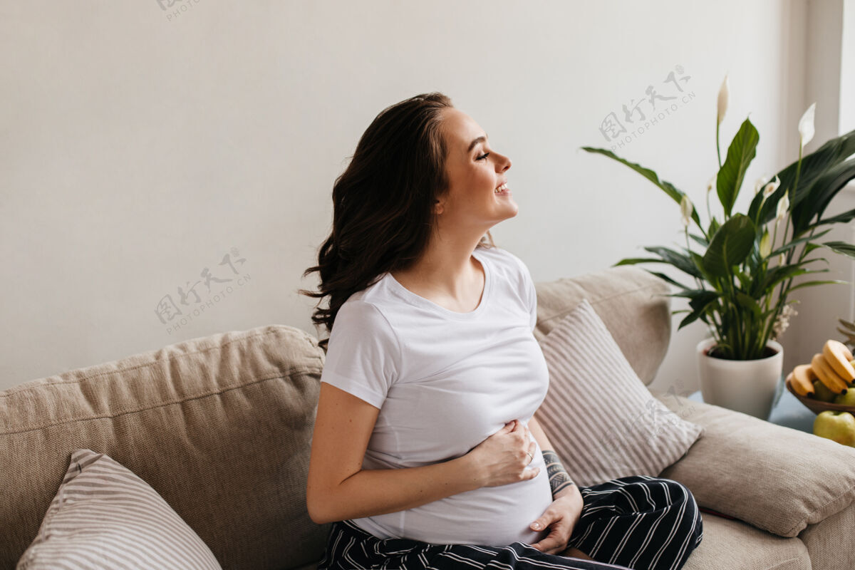 沙发漂亮的黑发卷发孕妇在条纹裤和白色T恤微笑坐着女人怀孕