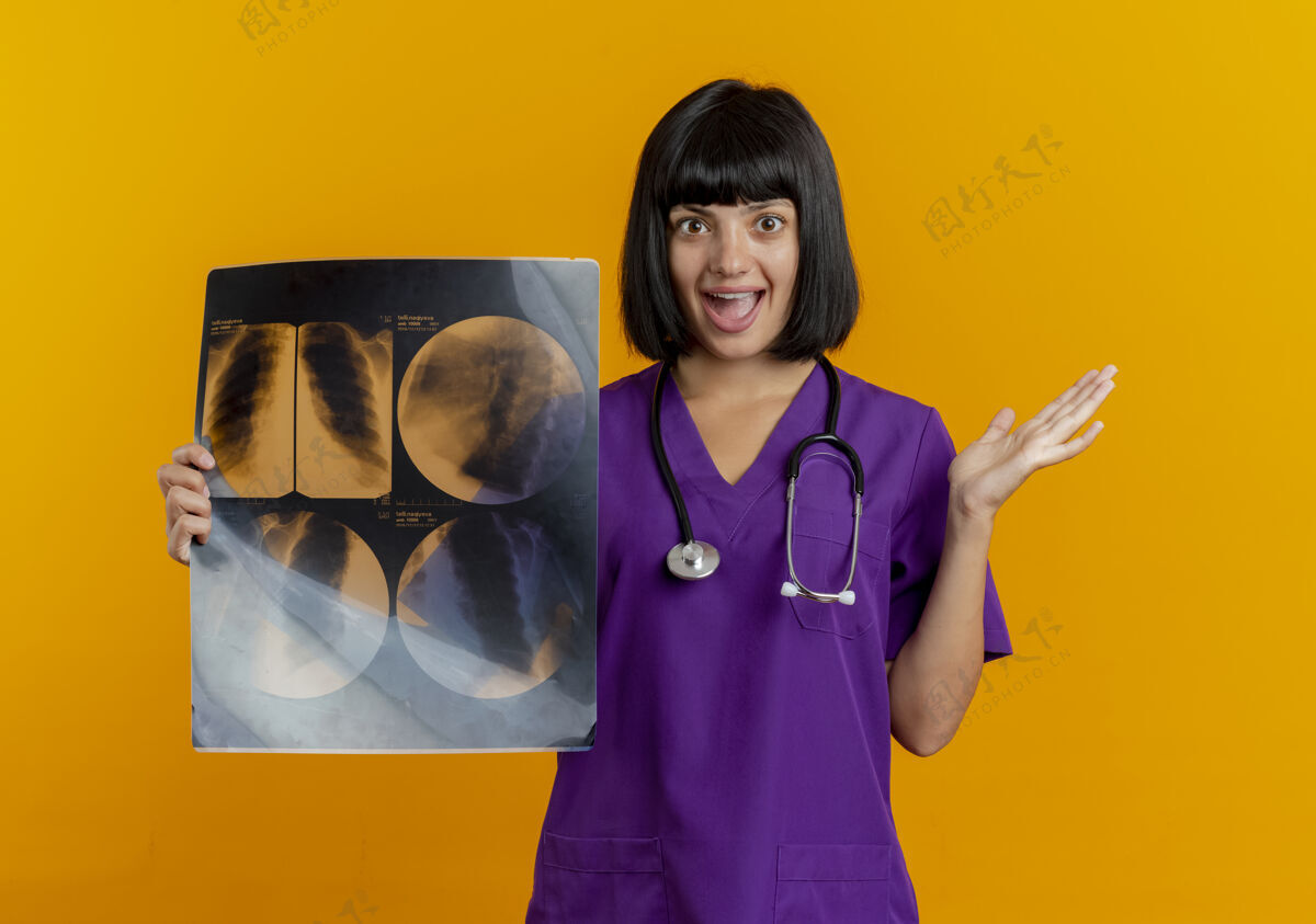 黑发兴奋的年轻黑发女医生穿着制服 手持听诊器 举起手拿着x光片 隔离在橙色背景和复印空间上手医生举起