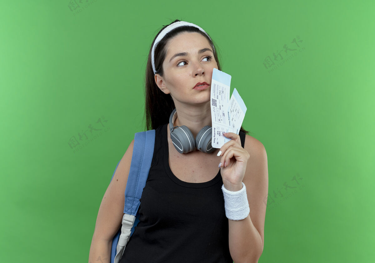 背包戴着头巾 背着背包 拿着机票 严肃的面孔站在绿色背景下的年轻健身女士空气严重面子