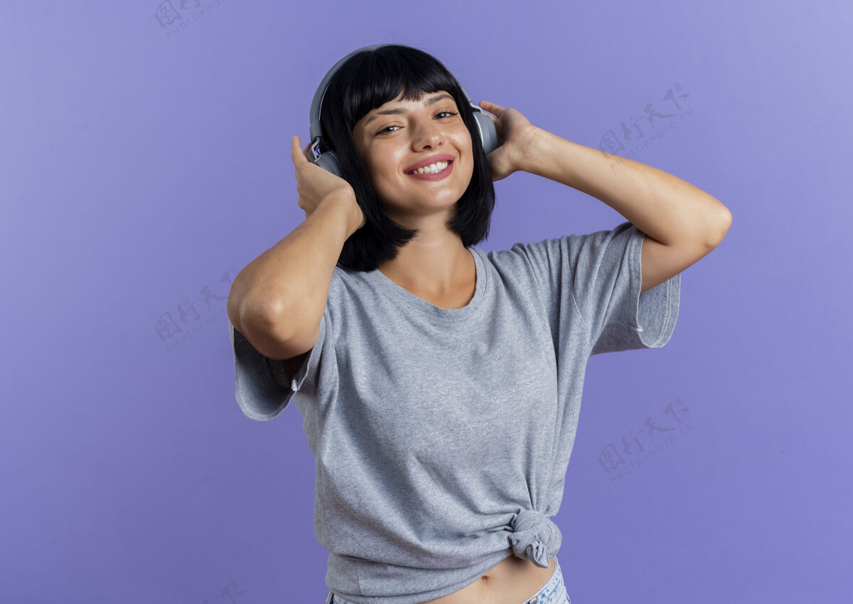 黑发戴着耳机的年轻黑发白人女孩微笑着看着隔离在紫色背景上的相机 并留有复印空间太空紫色微笑