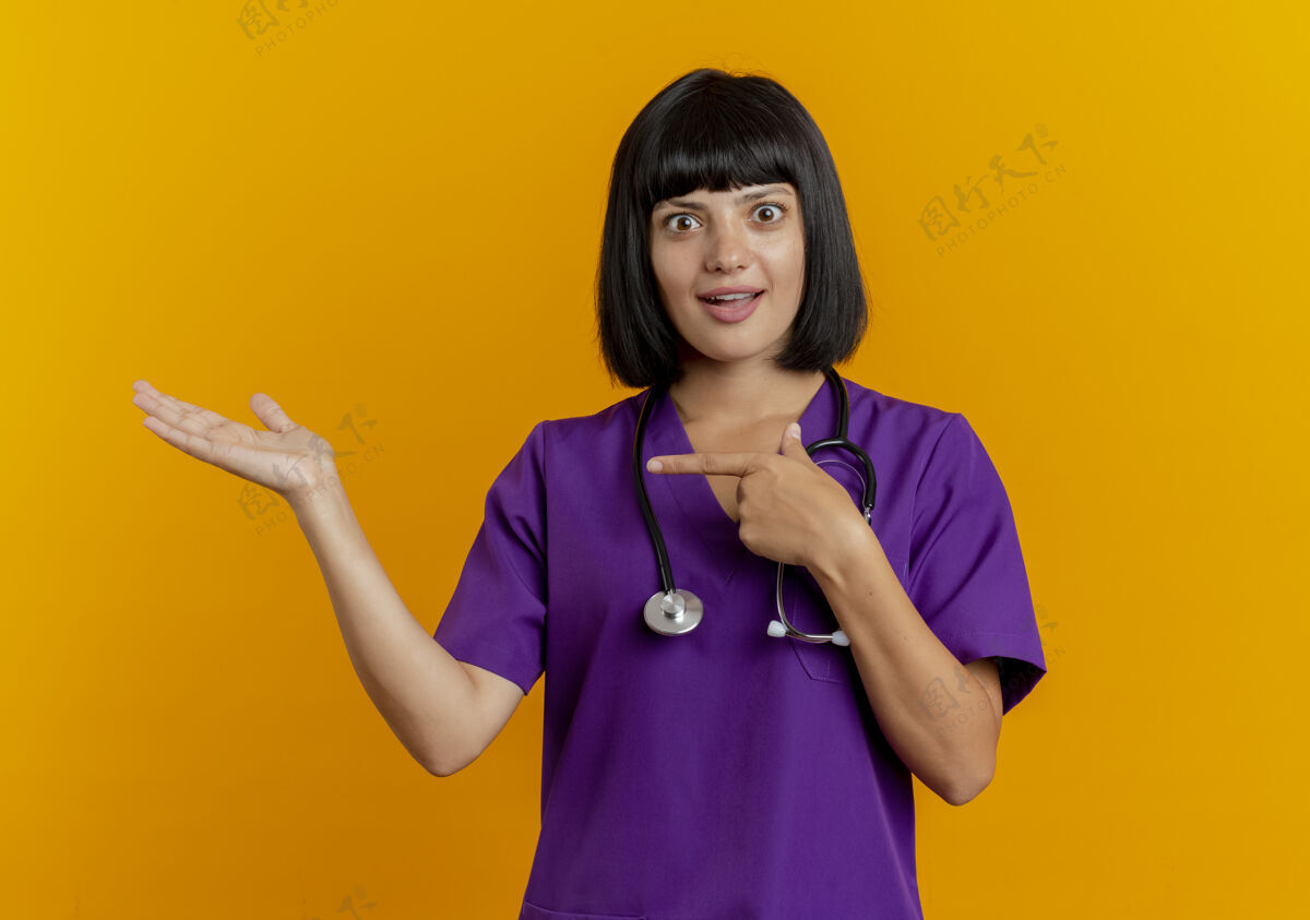 复制惊讶的年轻黑发女医生穿着制服 用听诊器指着被隔离在橙色背景上的空手 并留有复印空间空听诊器医生