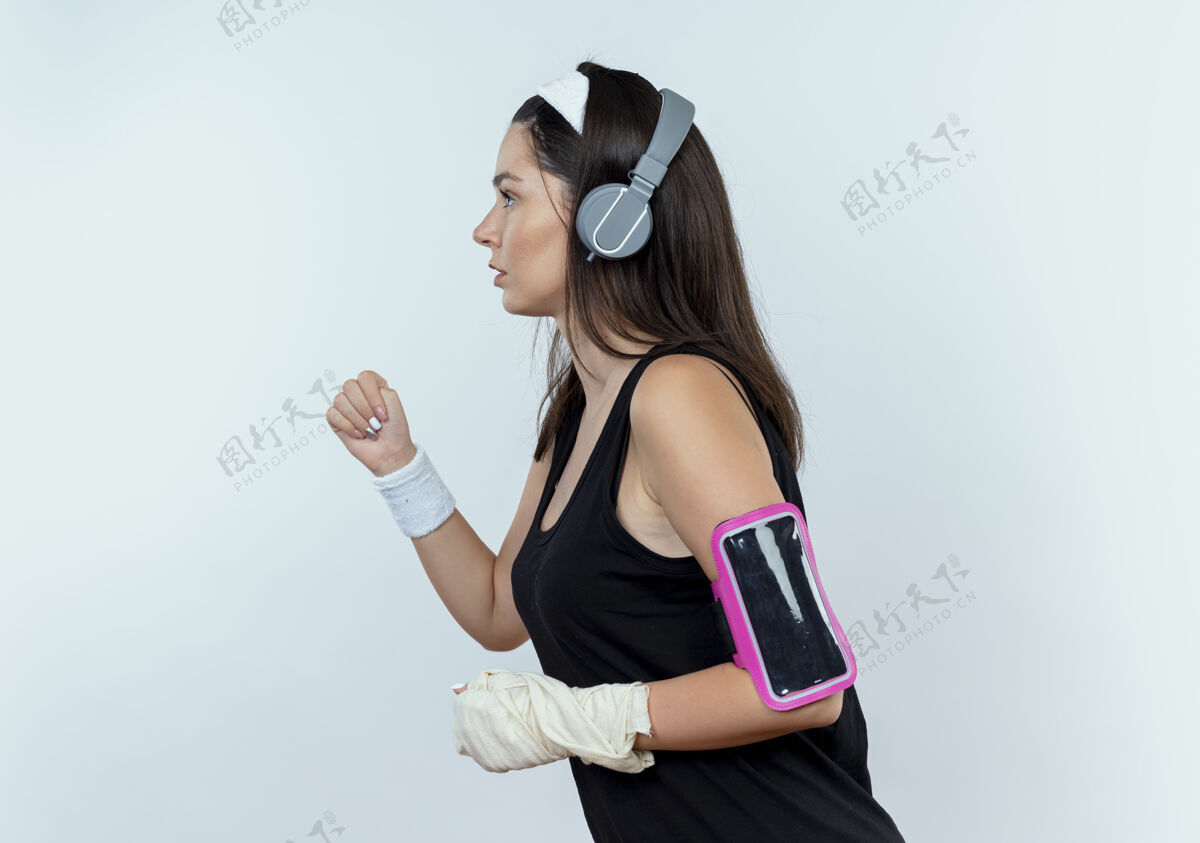 耳机戴着耳机和智能手机臂带的年轻健身女士站在白色背景下锻炼身体臂带手机站立