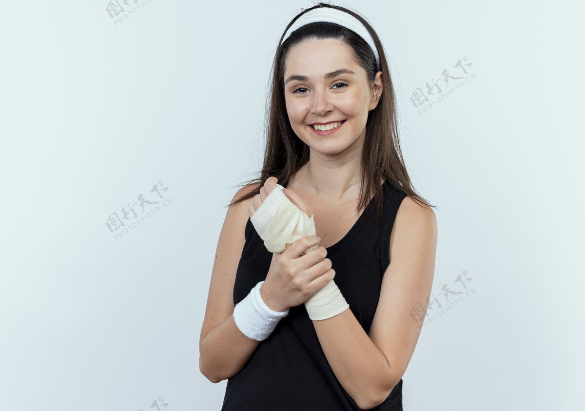 站立戴着头巾的年轻健身女士抚摸着她缠着绷带的手腕 站在白色背景上面带微笑地看着摄像机年轻相机面部