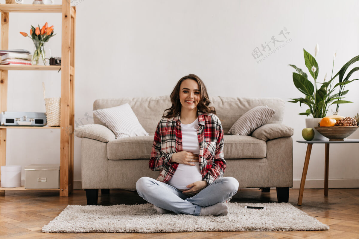 房间穿着格子衬衫和牛仔裤 妆容美丽的卷发孕妇坐在舒适的房间沙发旁柔软的米色地毯上 温柔地抚摸着腹部 真诚地微笑着快乐美丽腹部