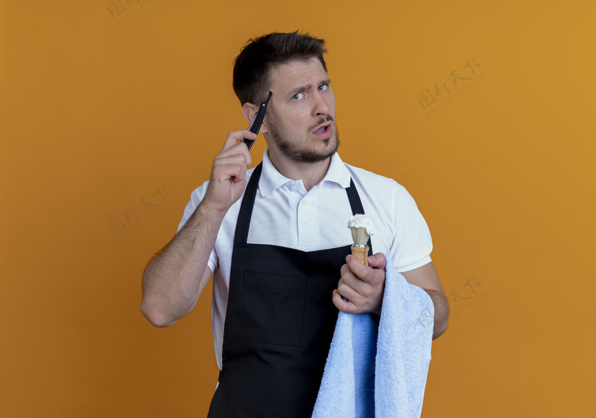 围裙围裙上的理发师手里拿着毛巾 手里拿着泡沫剃须刷和剃须刀 站在橙色背景上困惑地看着摄像机刷子泡沫拼图