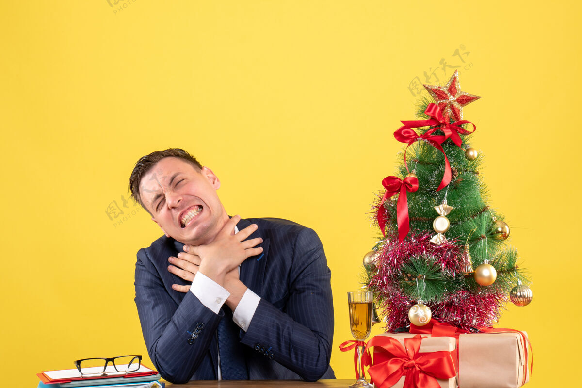 前面前视图愤怒的男子双手勒死自己坐在圣诞树和黄色背景上的礼物旁边的桌子上生意人桌子男人