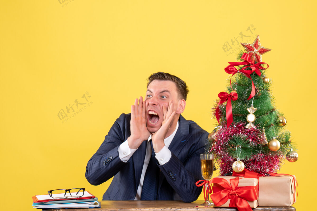 人前视图愤怒的男子一边大声喊叫 一边坐在圣诞树旁的桌子上 呈现黄色背景男人交流同时