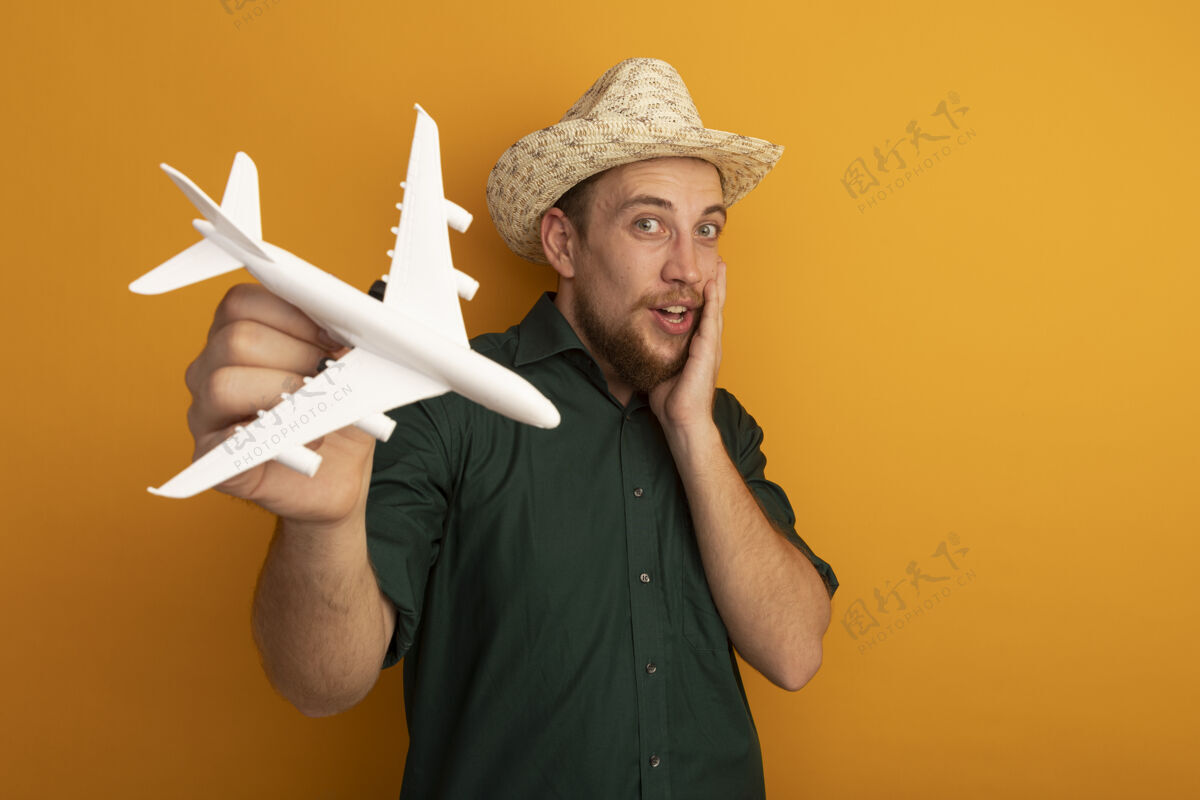 男人兴奋的金发帅哥 戴着沙滩帽 把飞机模型孤立地放在橙色的墙上帽子举行模特