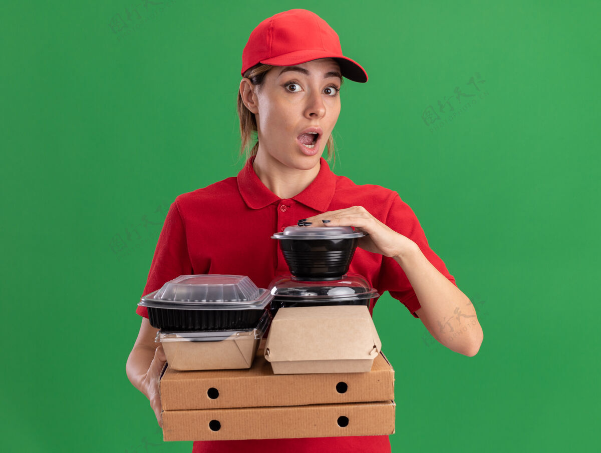 脸令人印象深刻的年轻漂亮的女送货员穿着制服拿着纸食品包装和比萨饼盒上的容器隔离在绿色的墙壁上盒子制服站着