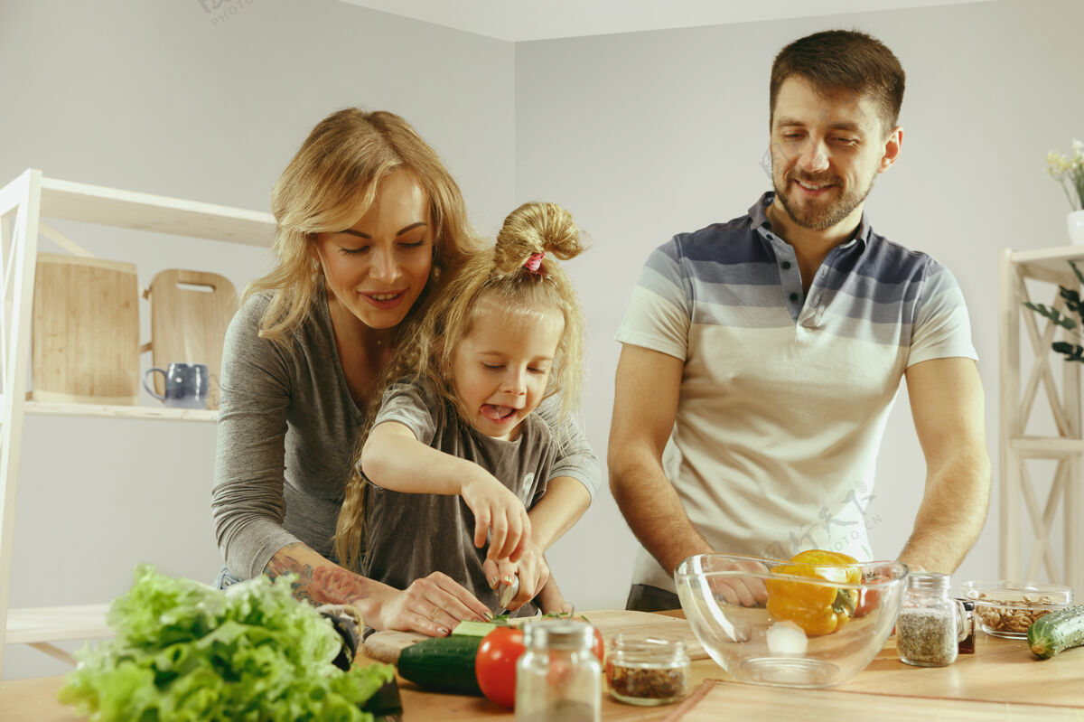 乐趣可爱的小女孩和她美丽的父母正在家里的厨房里切菜 一边微笑着做沙拉家庭生活理念年轻爸爸新鲜
