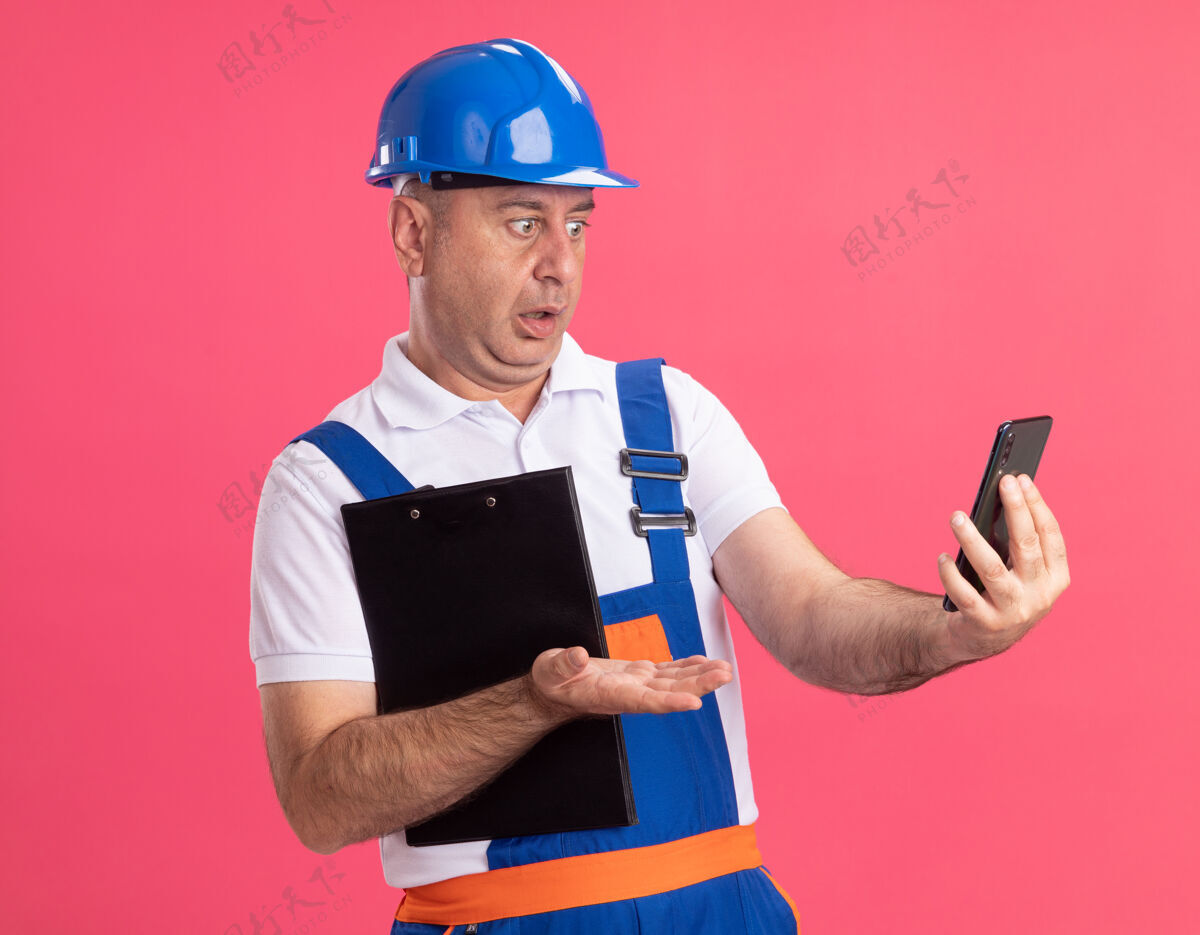 电话震惊的穿制服的成年建筑工人拿着剪贴板 看着隔离在粉红色墙上的电话成人人制服