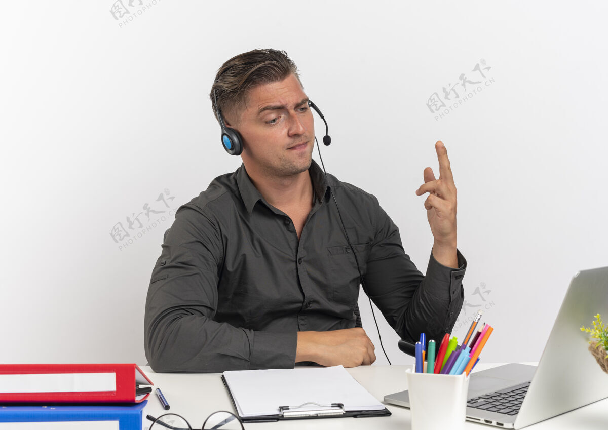 坐着年轻恼怒的金发上班族戴着耳机坐在办公桌旁 手里拿着办公工具 抬起手看着笔记本电脑 背景是白色的 还有复印空间手金发碧眼年轻人