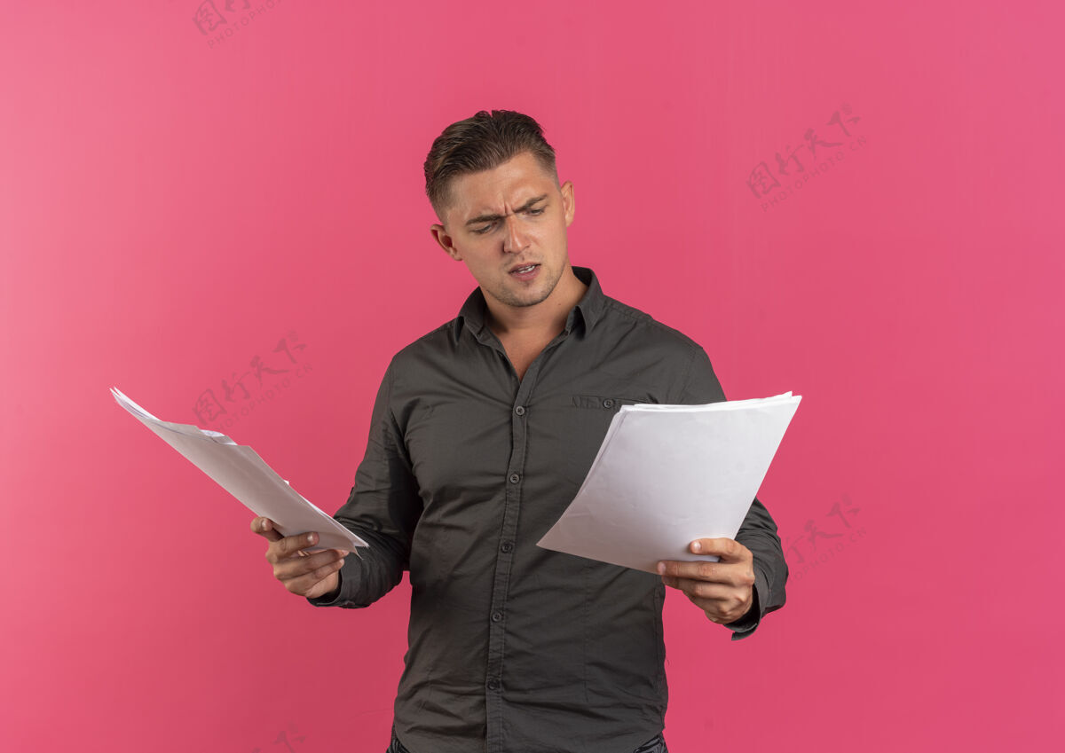 英俊年轻恼怒的金发帅哥拿着 看着粉色背景上有复印空间的纸页抱着生气年轻