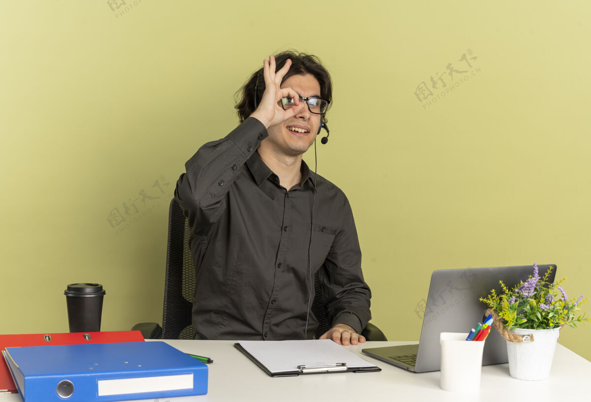 工具戴着耳机 戴着眼镜 面带微笑的年轻上班族坐在办公桌旁 手里拿着办公工具 手里拿着笔记本电脑 隔着手指在绿色背景上看 还有复印空间微笑年轻办公桌