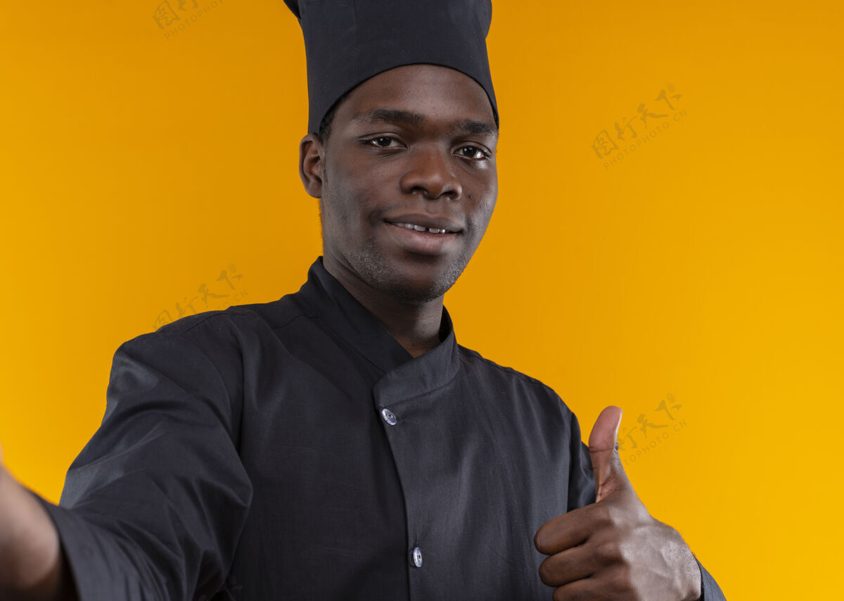 假装身着厨师制服 面带微笑的年轻美国黑人厨师假装拿着相机 竖起大拇指孤立地站在橙色背景上 留着复印空间非裔美国人相机拿着