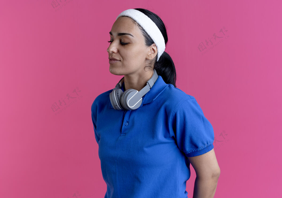 自信年轻自信的高加索运动女性戴着头带和腕带站着 脖子上戴着耳机 粉色背景上有复制空间耳机颈部运动