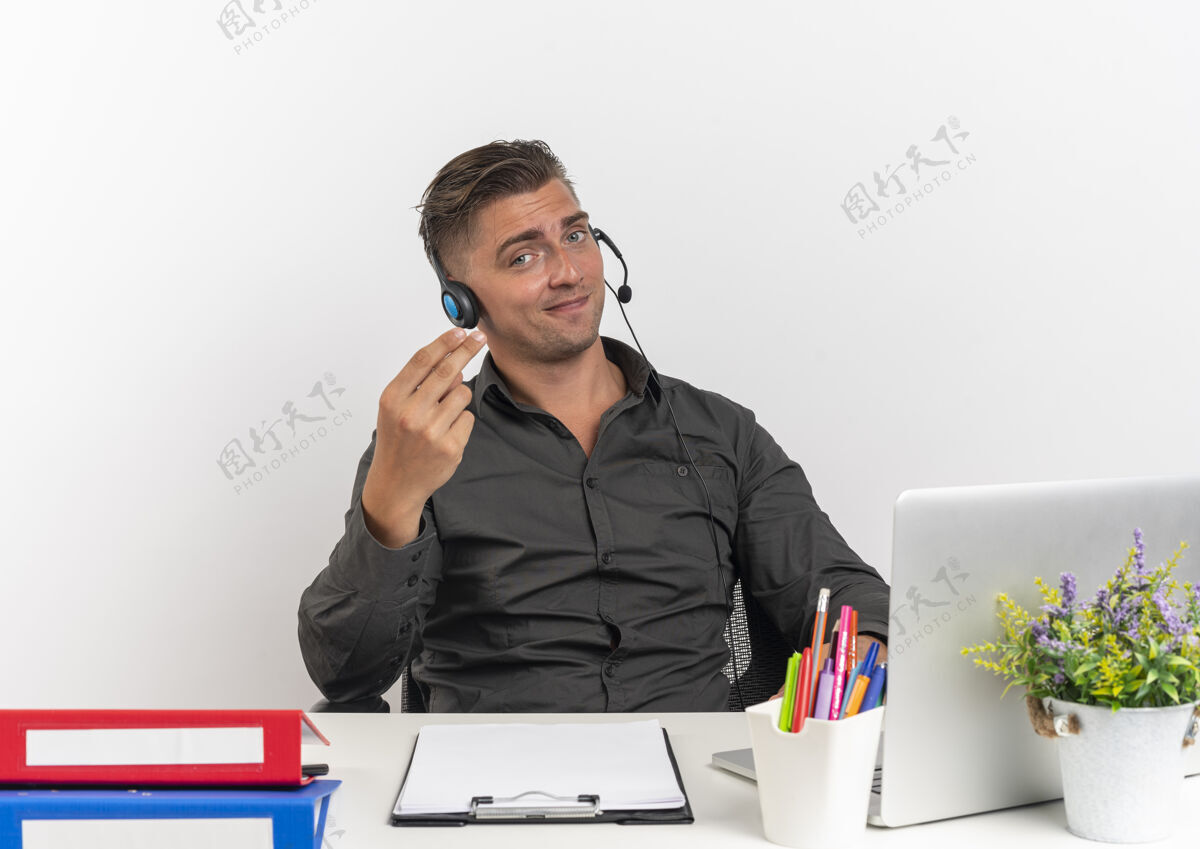 工人年轻而快乐的金发上班族戴着耳机坐在办公桌旁 拿着办公工具 用笔记本电脑做手势 在白色背景上孤立着钱的手势 还有复印空间钱年轻金发