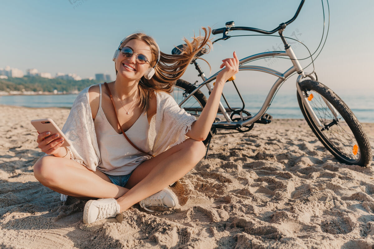 休闲时尚年轻迷人的金发微笑的女人坐在沙滩上骑着自行车戴着耳机听音乐耳机活跃沙滩