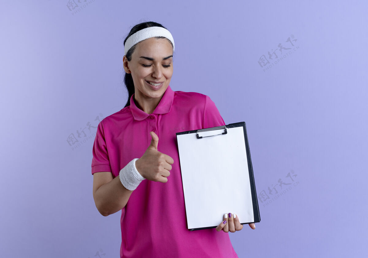 年轻年轻微笑的白人运动女性戴着头带和腕带大拇指举着剪贴板在紫色背景上与复制空间隔离头带微笑运动