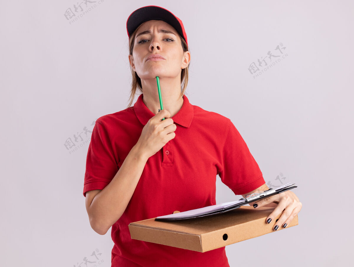 站着自信的年轻漂亮的女送货员穿着制服 把铅笔放在下巴上 把剪贴板放在隔离在白墙上的比萨饼盒上公民人脸