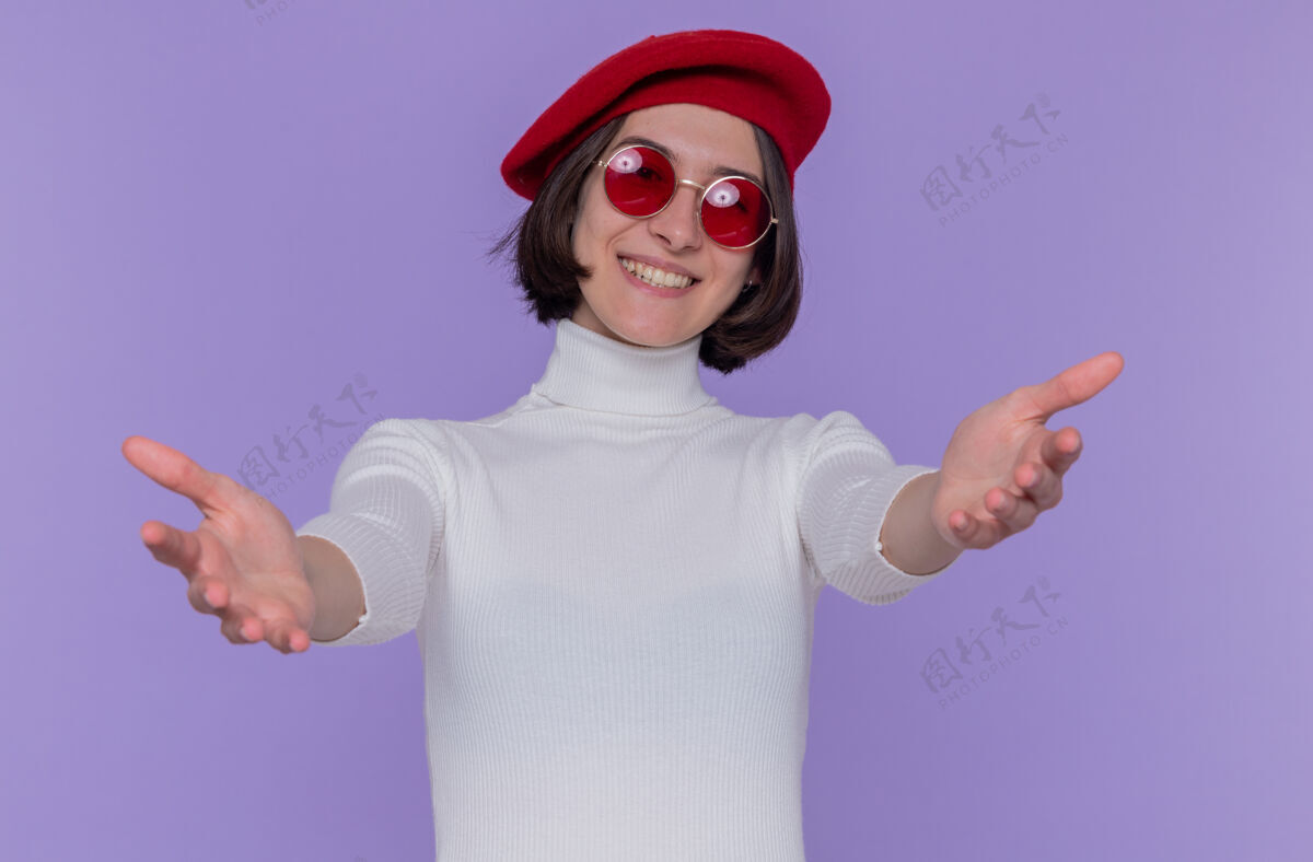 手快乐而积极的年轻女子 短发 白色高领毛衣 头戴贝雷帽 戴着红色太阳镜来微笑年轻