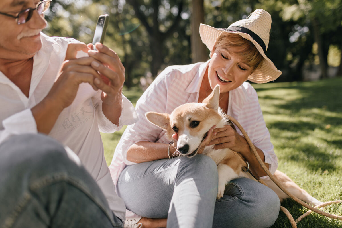 欢乐戴着帽子 穿着粉色衬衫 留着短发的时髦女士微笑着 坐在草地上 和公园里的科吉和拿着智能手机的男人合影自然模特帅哥