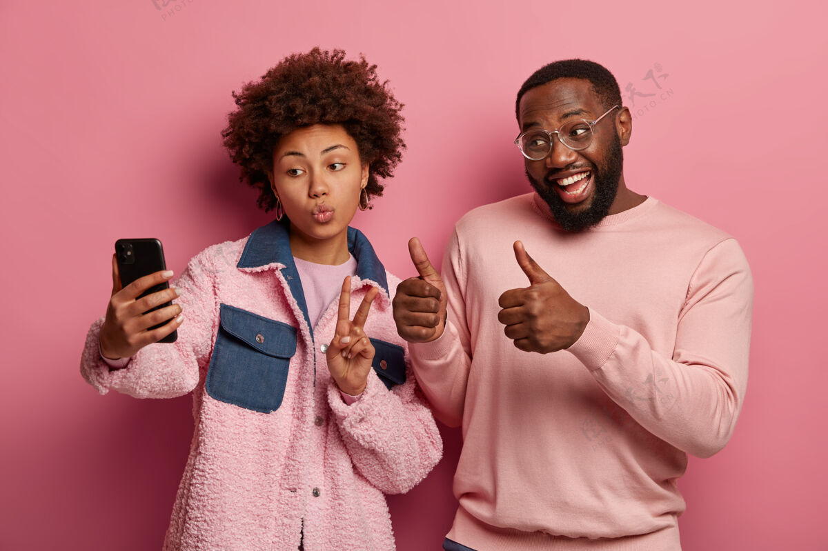 和平照片中的黑人男女在手机上自拍肖像 做出和平和喜欢的手势 积极地看着智能手机相机 穿粉红色的衣服高兴请自拍
