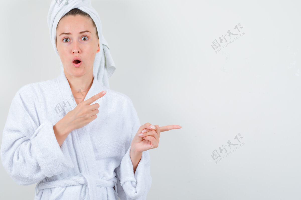 漂亮年轻的女士指着旁边的白色浴衣 毛巾 看着惊讶前视图自然成人女士