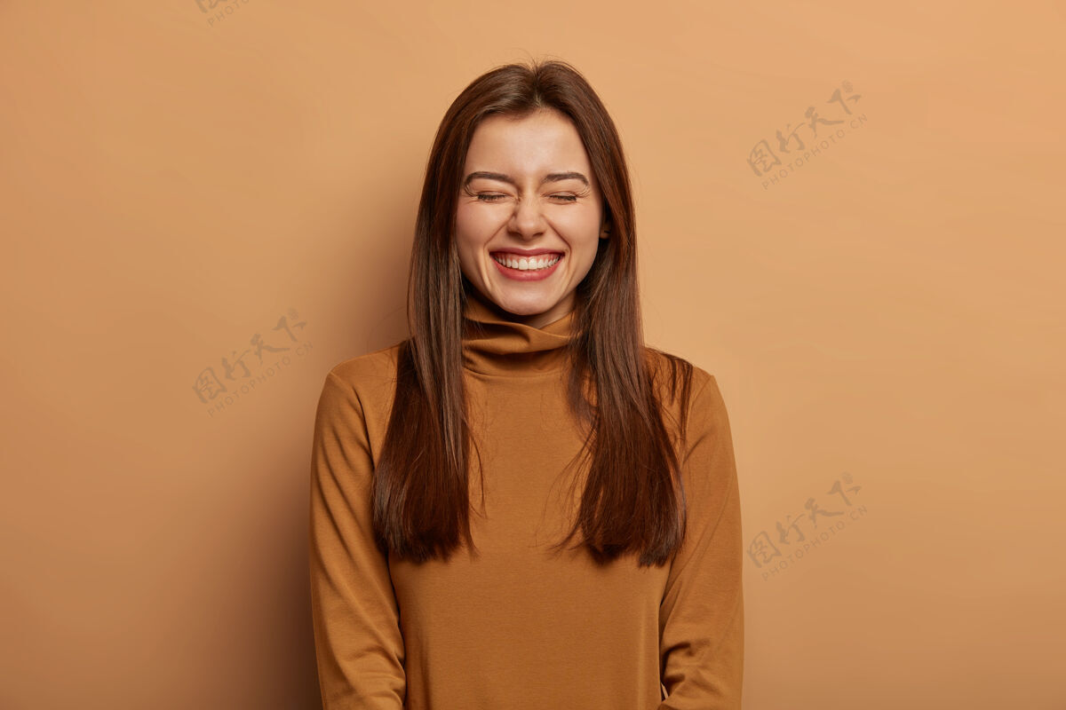 情绪俏皮好笑的女人留着长长的深色直发 欢笑着 穿着棕色的领毛衣 开心地笑着高兴姿势请
