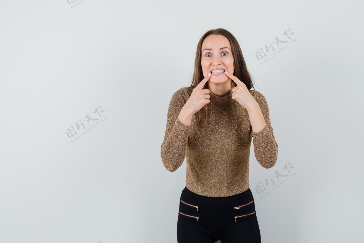 手指年轻女子穿着金色的镀金毛衣和黑色的裤子 把食指放在嘴边 强颜欢笑 看起来很开心 前视力量护理脸