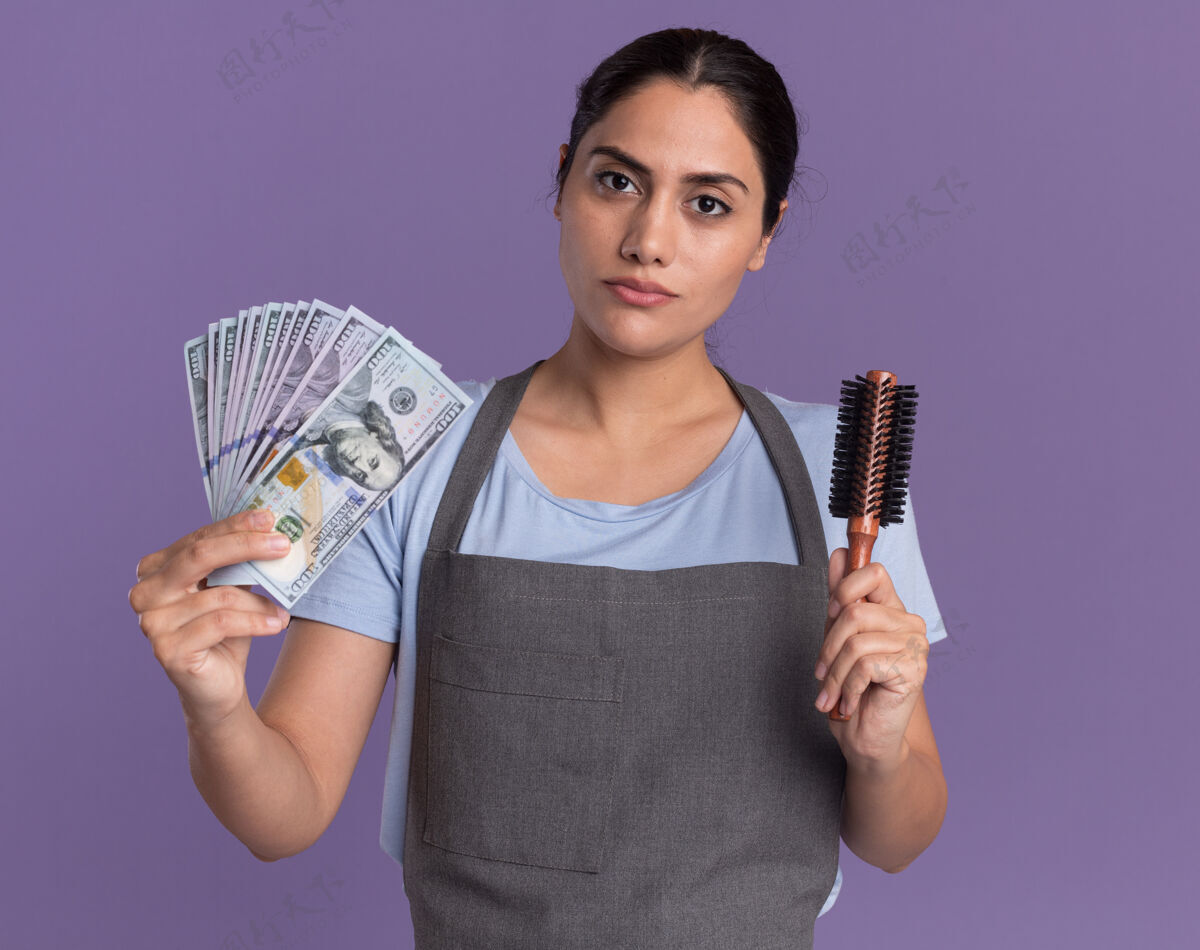 严肃穿着围裙的年轻漂亮的女理发师拿着现金和发刷 严肃的脸站在紫色的墙上看着前面漂亮抱着现金