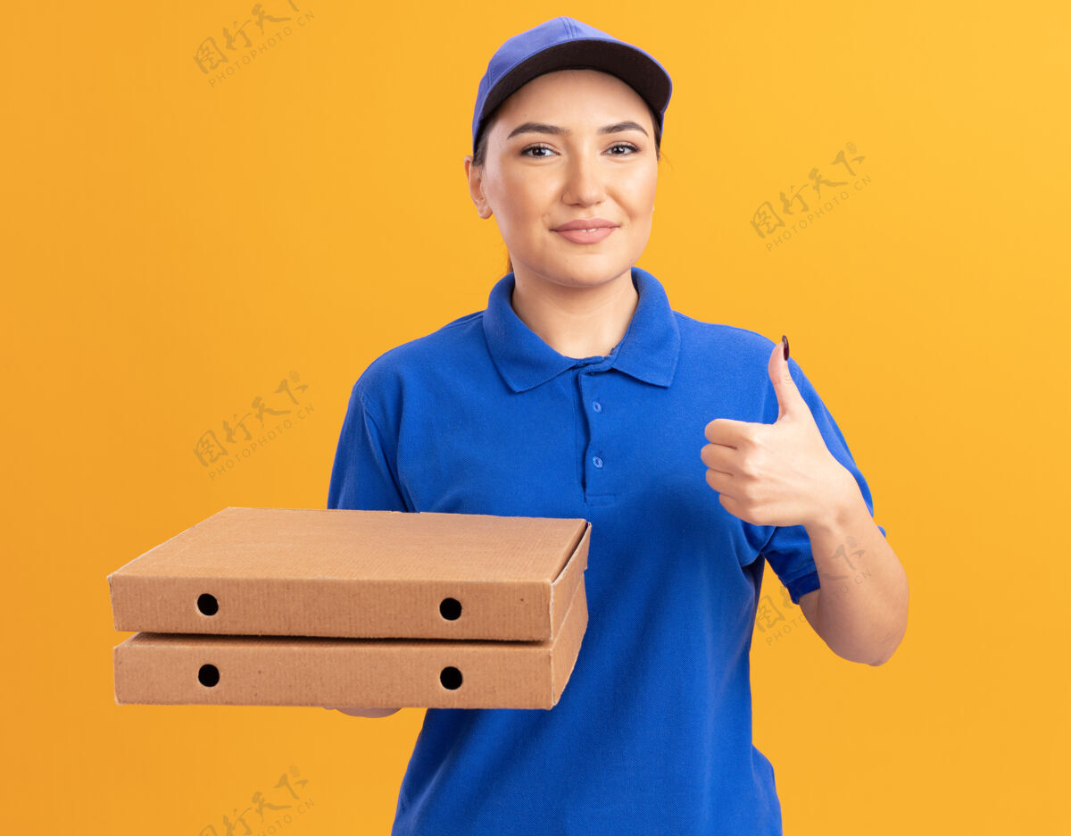 站着快乐的年轻送货员身穿蓝色制服 戴着帽子 拿着披萨盒 微笑自信地站在橙色的墙上竖起大拇指 看着前面盒子微笑展示