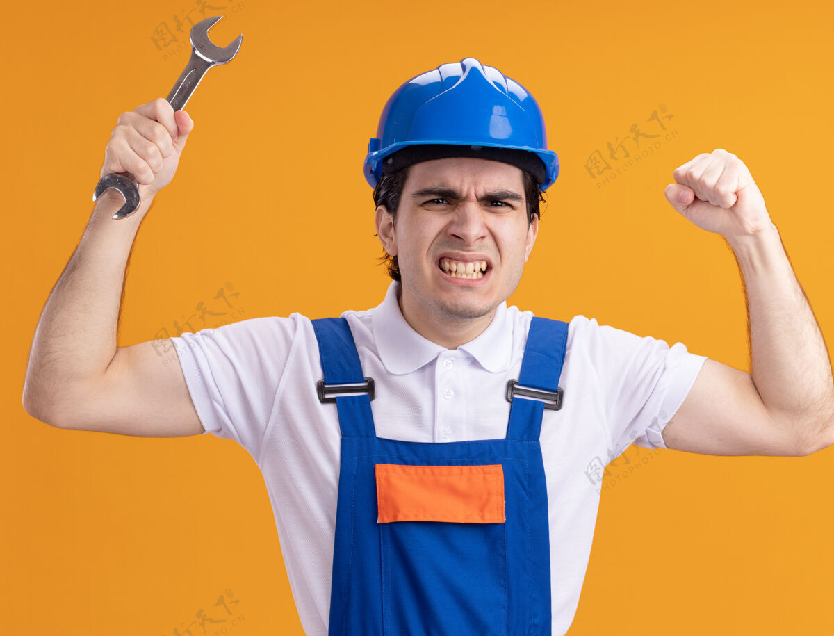 扳手年轻的建筑工人穿着建筑制服 戴着安全帽 手里拿着扳手 站在橘色的墙上生气又不高兴地举手拿着举起安全