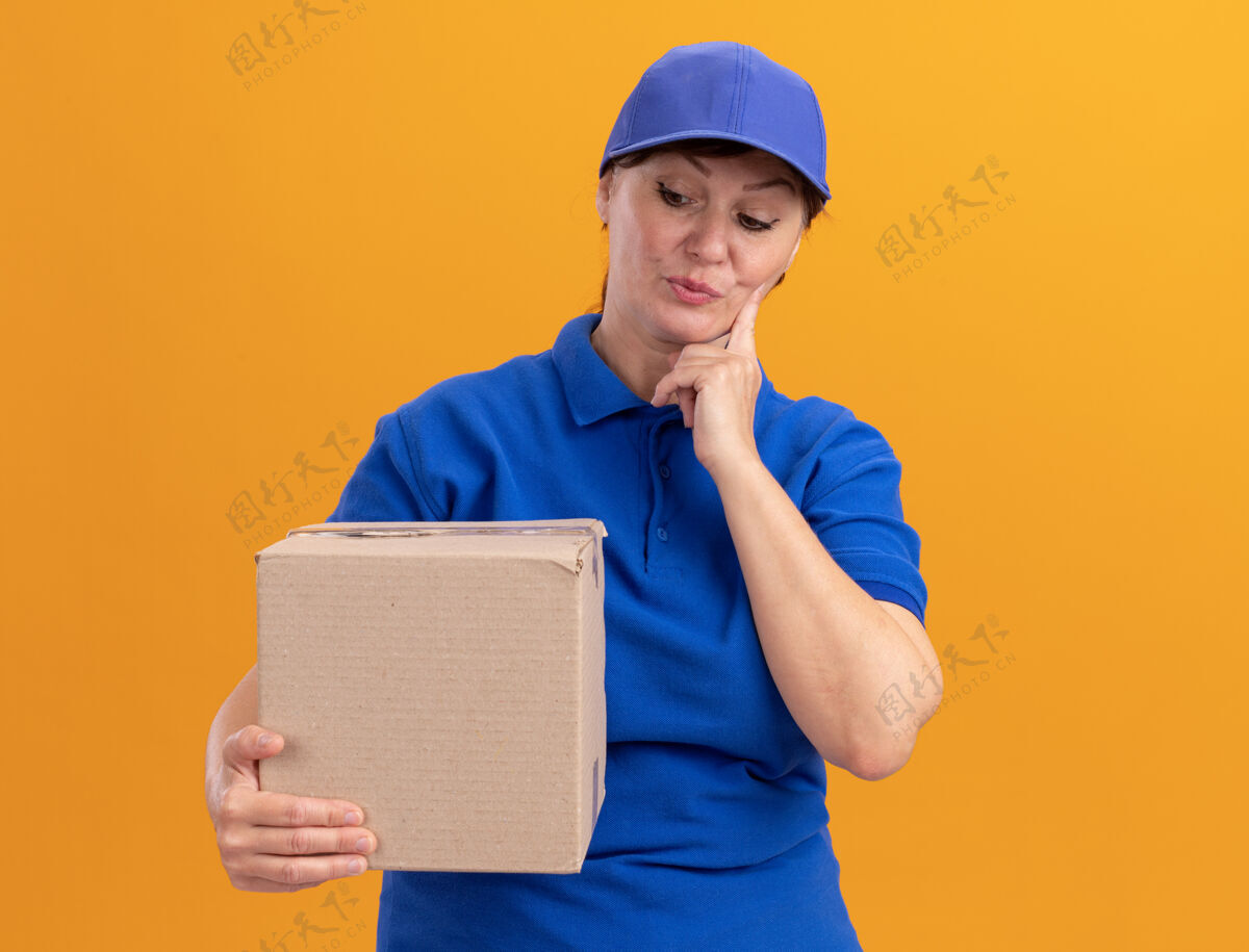 拼图中年送货员身穿蓝色制服 戴着帽子 手里拿着纸板箱 站在橘色的墙上困惑地看着它拿着盒子女人