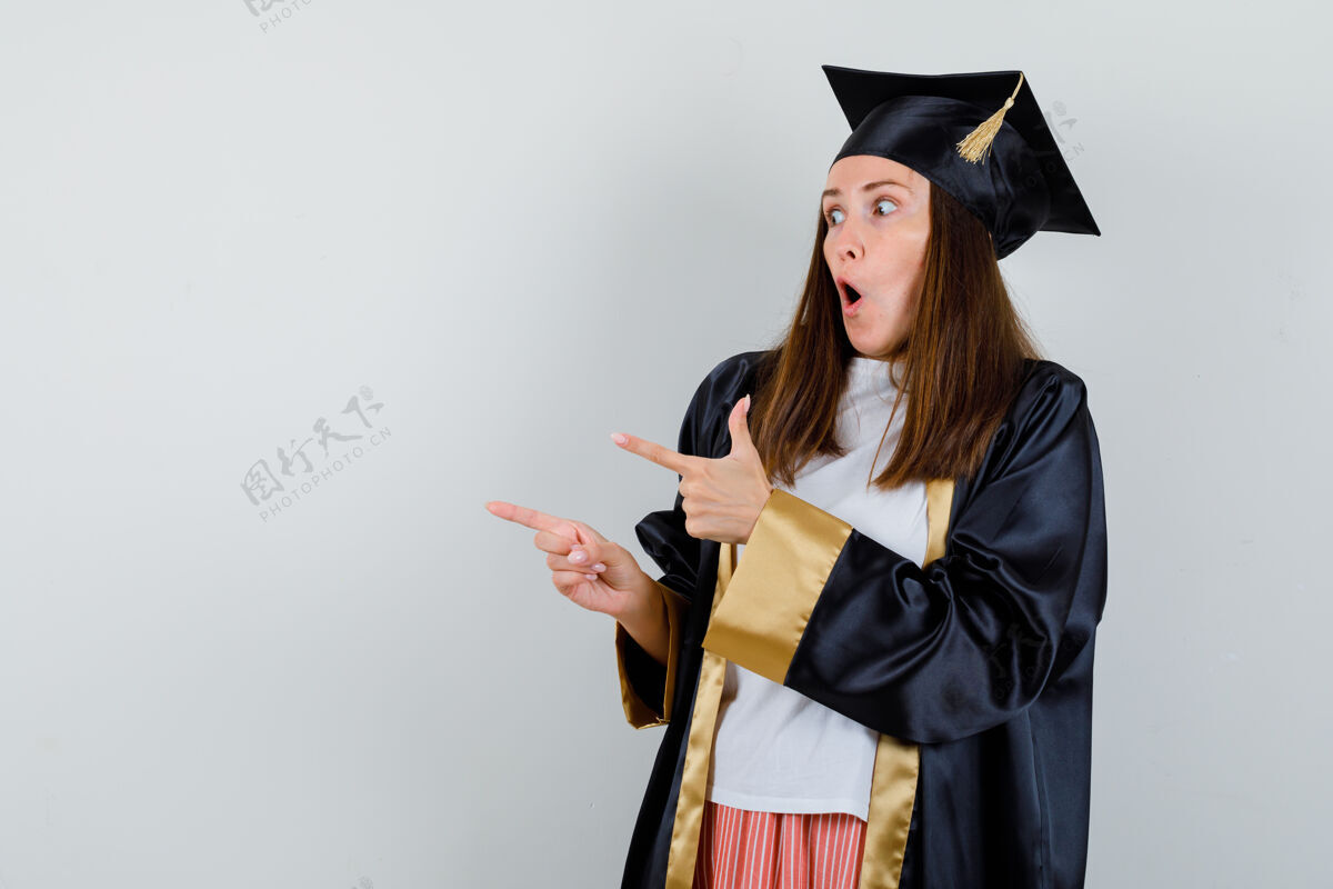 成功毕业妇女的肖像指着旁边的休闲服 制服和期待惊讶的前视图女性长袍学院