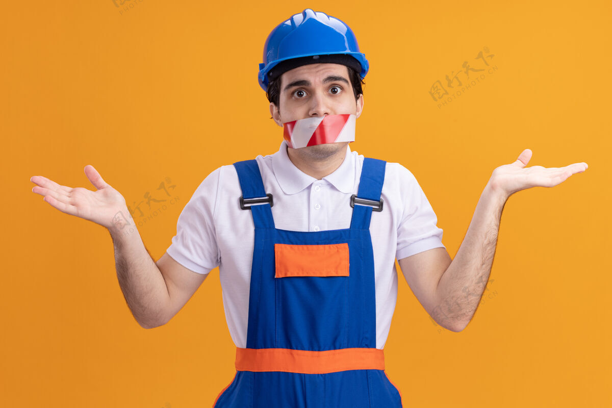 建筑年轻的建筑工人穿着建筑制服 戴着安全帽 嘴上缠着胶带 困惑地耸耸肩站在橙色的墙上围着困惑裹着
