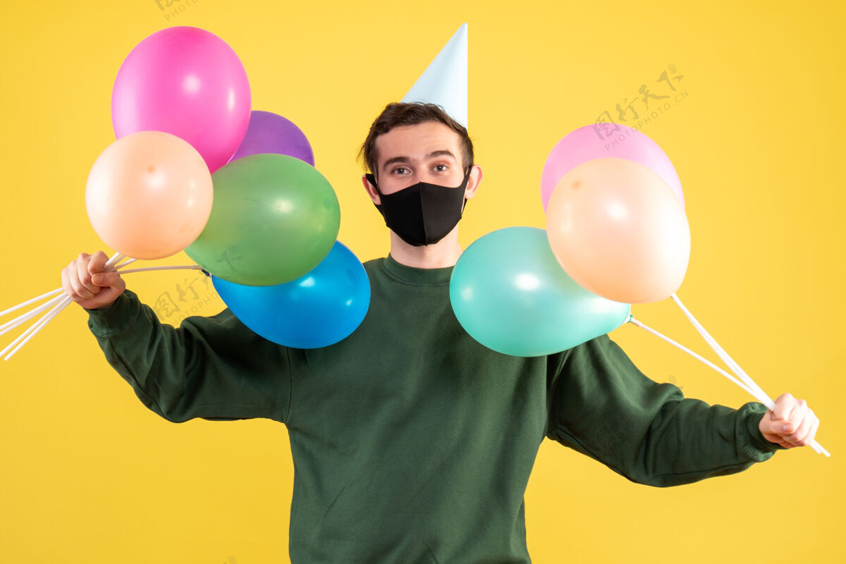 立场正面图：戴着派对帽的年轻人双手捧着气球站在黄色背景上气球乐趣手